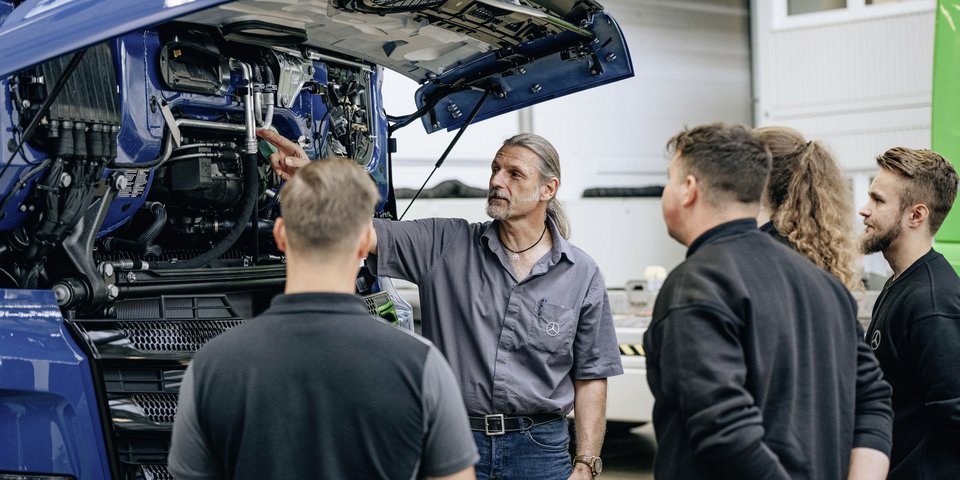 An instructor explains an open engine compartment of a blue Daimler truck to four trainees.