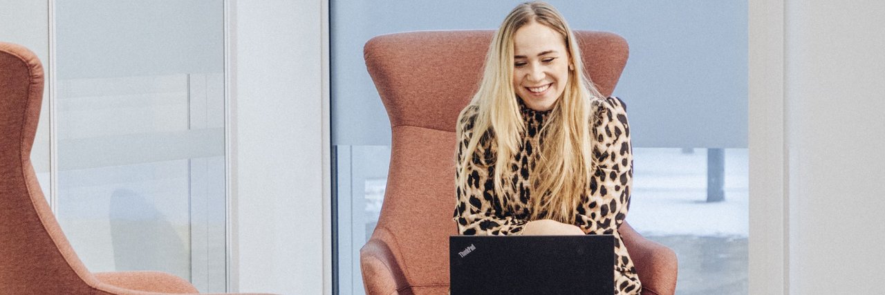 Eine junge Mitarbeiterin sitzt in einem hellen, modernen Lounge-Bereich auf einem Sessel und arbeitet am Laptop auf einem kleinen Tisch vor ihr.