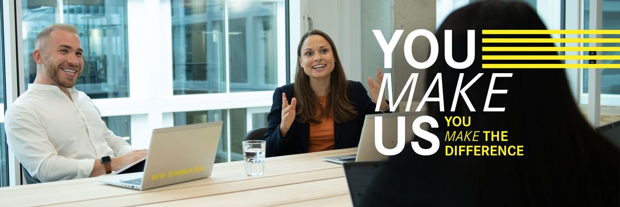 Drei Kollegen sitzen sich lachend im Büro an einem Besprechungstisch mit Laptops gegenüber. Über dem Bild liegt der "You Make Us" Schriftzug.
