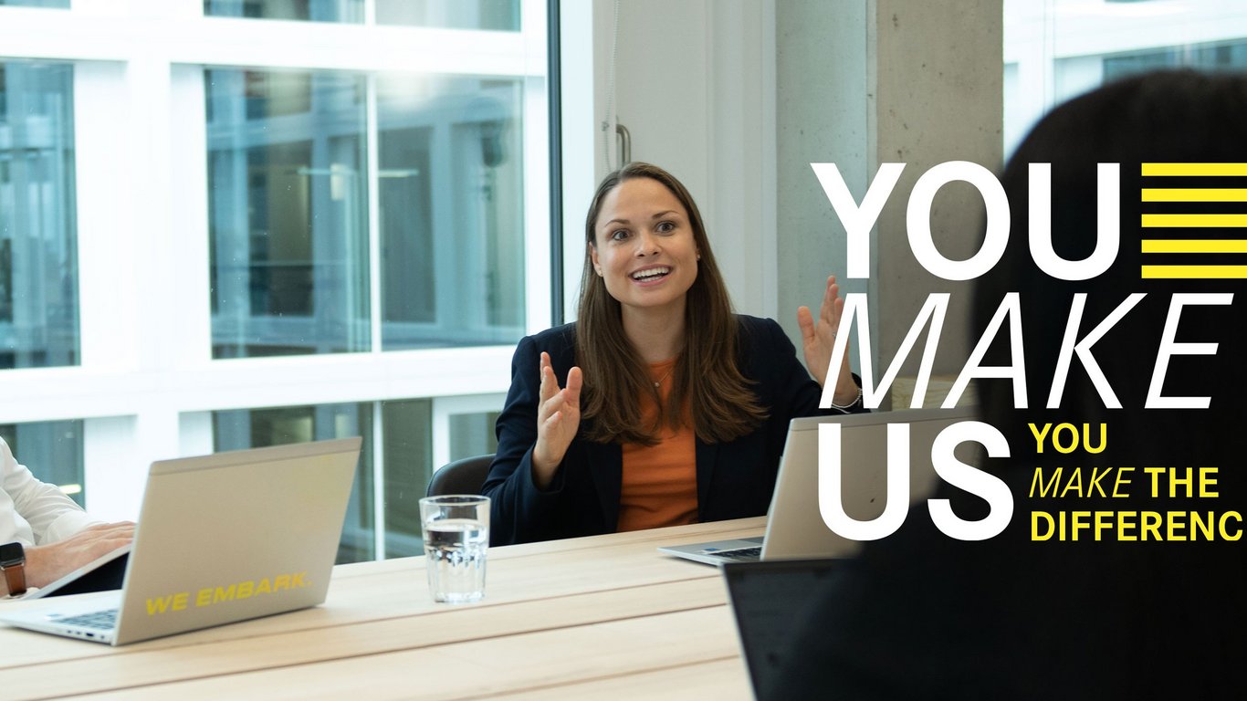 Drei Kollegen sitzen sich lachend im Büro an einem Besprechungstisch mit Laptops gegenüber. Über dem Bild liegt der "You Make Us" Schriftzug.