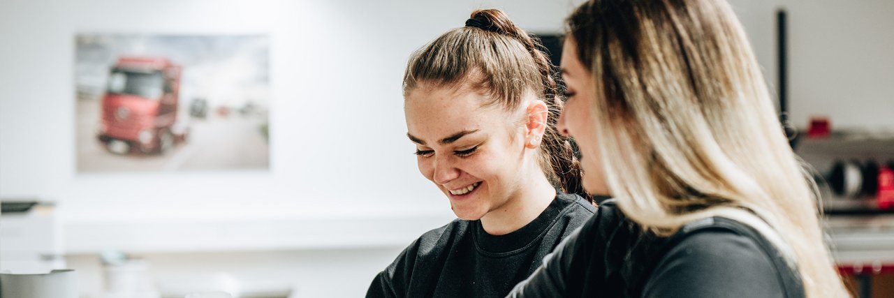 Zwei fröhliche Auszubildende in schwarzer Arbeitskleidung verpacken gemeinsam eine Kiste.