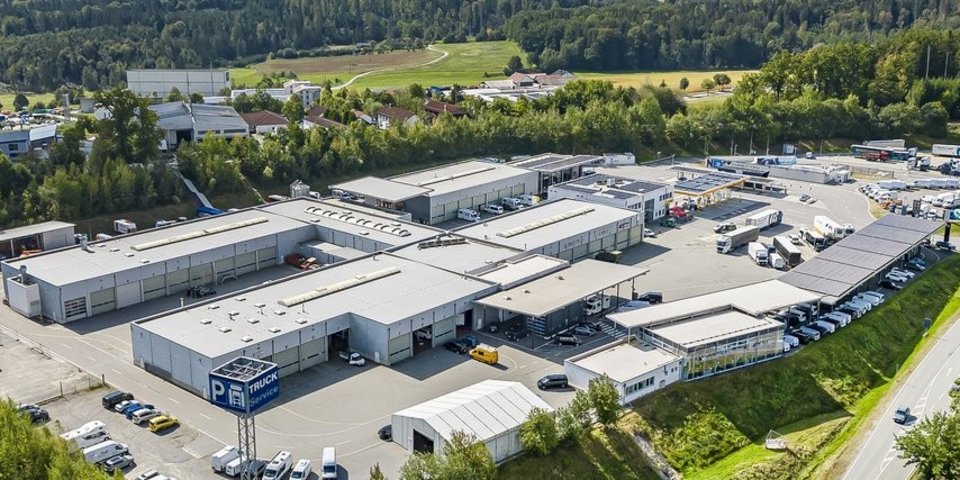 Luftaufnahme des Nutzfahrzeugzentrums Daimler Truck in Passau mit mehreren großen Werkshallen, Parkplätzen für Lkw und Pkw sowie angrenzender Straße und grüner Landschaft im Hintergrund.