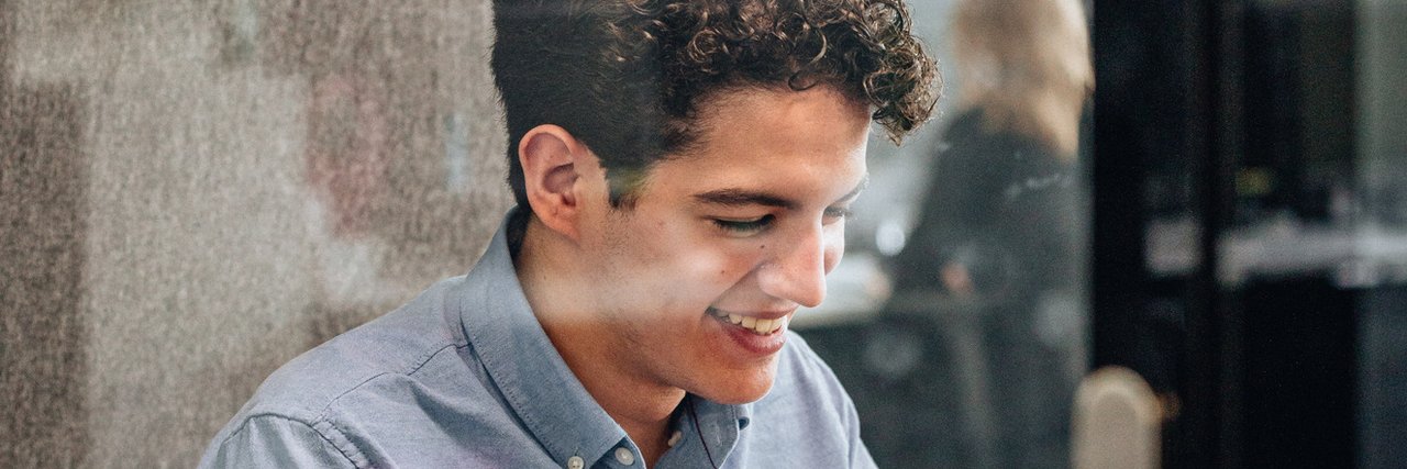 Ein junger kaufmännischer Auszubildender arbeitet lächelnd in einem verglasten Meetingraum an seinem Laptop.