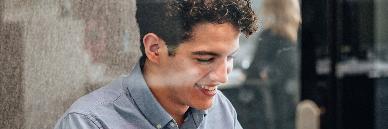 Ein junger kaufmännischer Auszubildender arbeitet lächelnd in einem verglasten Meetingraum an seinem Laptop.