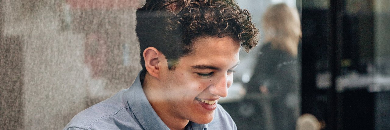 Ein junger kaufmännischer Auszubildender arbeitet lächelnd in einem verglasten Meetingraum an seinem Laptop.