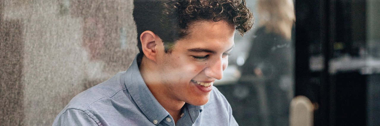 Ein junger kaufmännischer Auszubildender arbeitet lächelnd in einem verglasten Meetingraum an seinem Laptop.