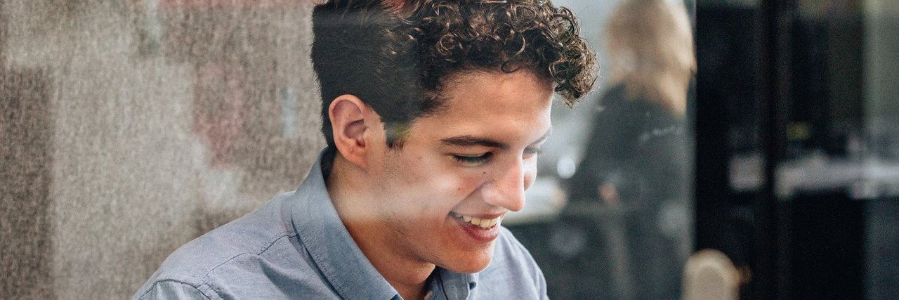 Ein junger kaufmännischer Auszubildender arbeitet lächelnd in einem verglasten Meetingraum an seinem Laptop.