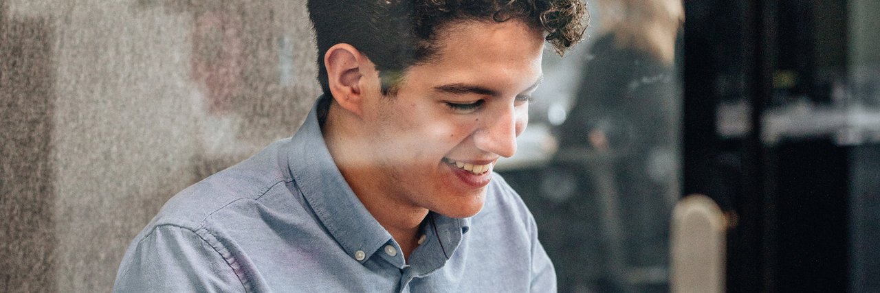 A young commercial trainee smiles as he works on his laptop in a glazed meeting room.