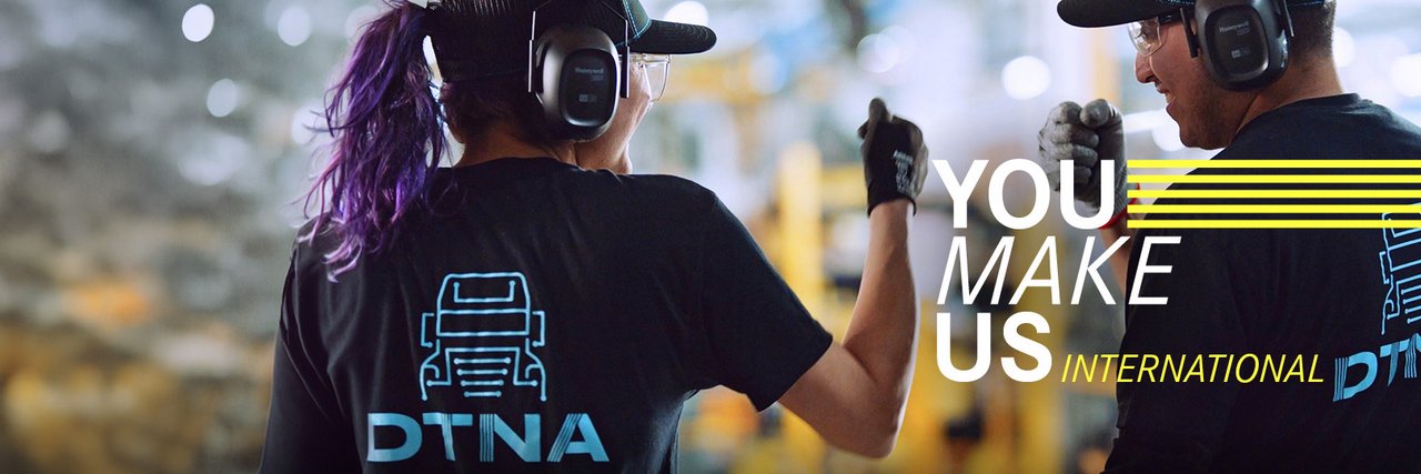 Zwei Mitarbeitende von Daimler Truck in einer Produktionsumgebung tragen Schutzkleidung und Headsets und geben sich ein motivierendes Handzeichen. Im Hintergrund ist eine unscharfe Fabrikhalle zu sehen. Der eingeblendete Slogan „You make us international“ betont die Bedeutung einer globalen Belegschaft.
