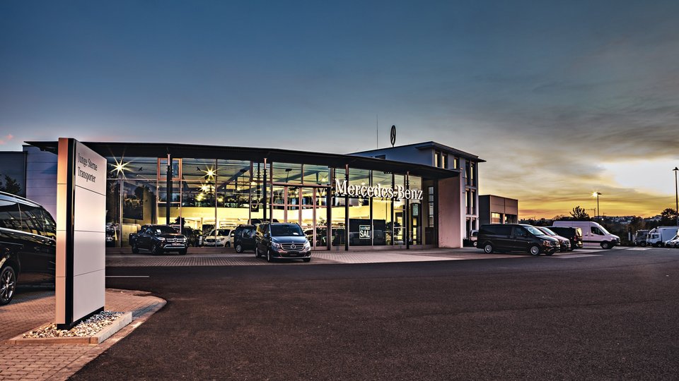 Mercedes-Benz Nutzfahrzeugzentrum Würzburg bei Sonnenuntergang, mit beleuchtetem Showroom und geparkten Transportern im Vordergrund.