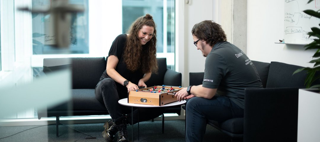Two employees sit on a sofa in the Daimler Truck Campus and play mini table football together.