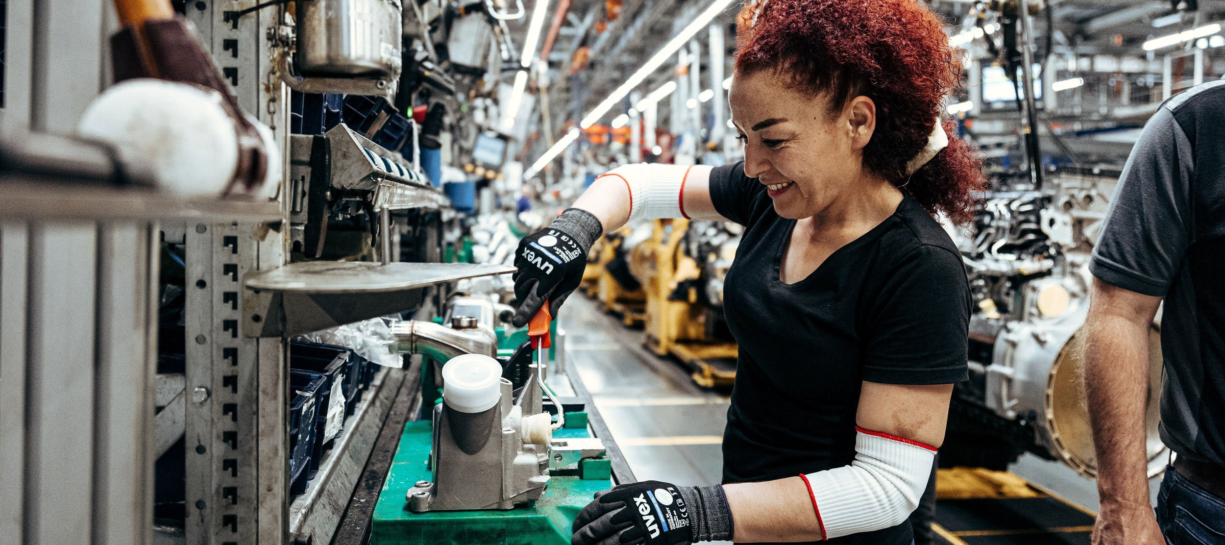 A red-haired woman wearing safety gloves works on a Daimler truck production part in a production hall.