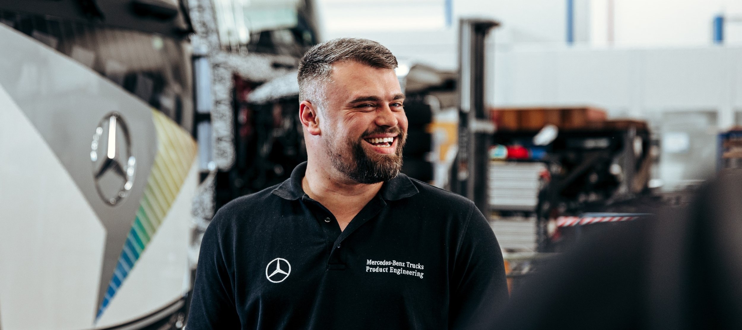 Employee in a black polo shirt smiles in a Mercedes-Benz Trucks workshop. A Daimler Truck lorry can be seen in the background.