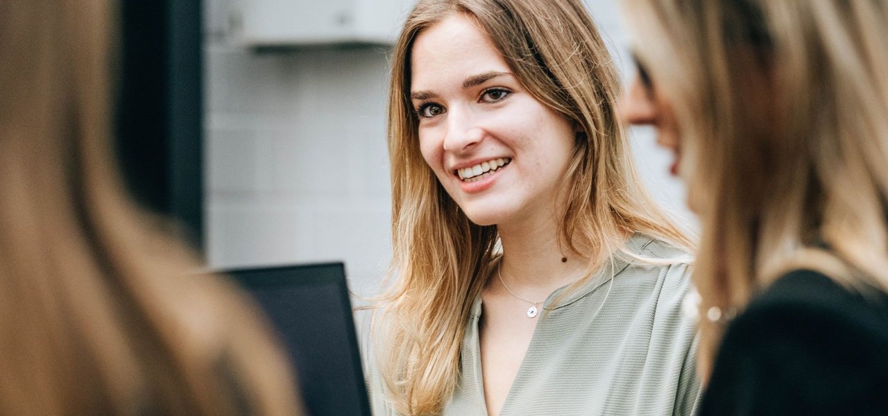 A young woman smiles and looks round as she sits with colleagues at a table with a laptop.