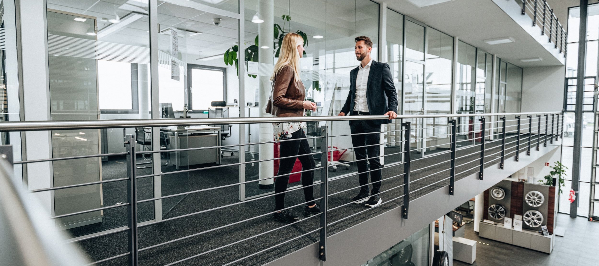 Two employees are chatting in a corridor with glass walls in Daimler Truck's modern Own Retail office building, surrounded by offices and sales areas.