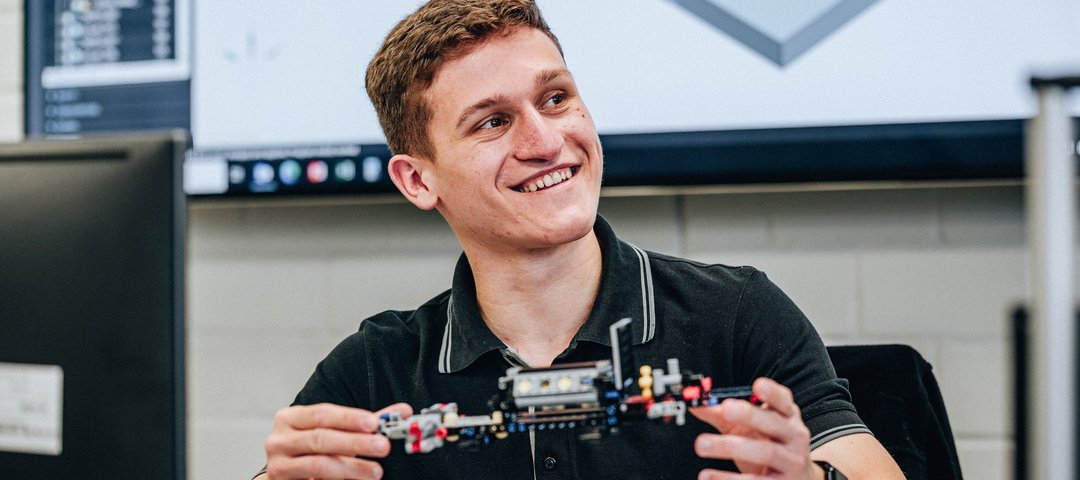A dual student smiles as he shows a technical model made of building blocks, with a monitor with CAD software in the background.