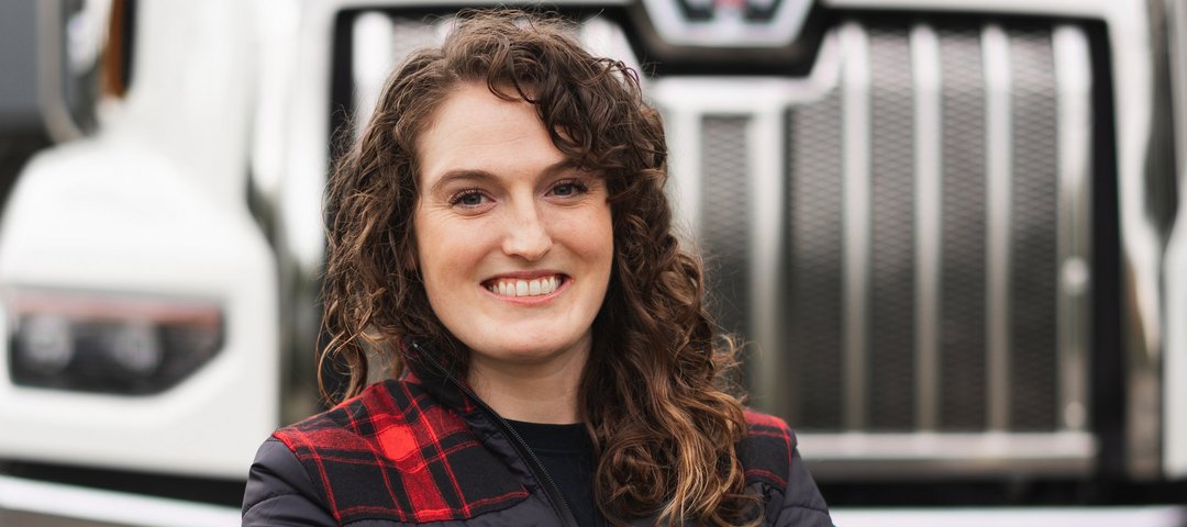 Portrait of Alex Banzer, a smiling woman with curly hair and folded arms stands in front of a large white truck.
