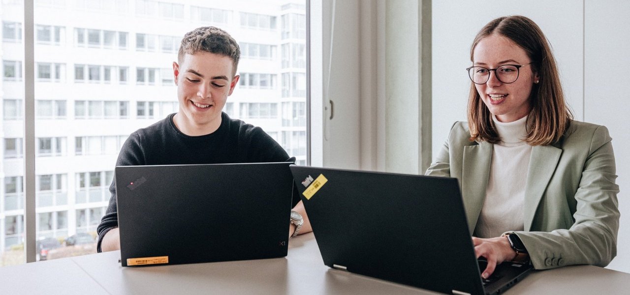 Zwei Doktoranten, ein junger Mann und eine junge Frau, sitzen nebeneinander und arbeiten am Laptop im Daimler Truck Campus.