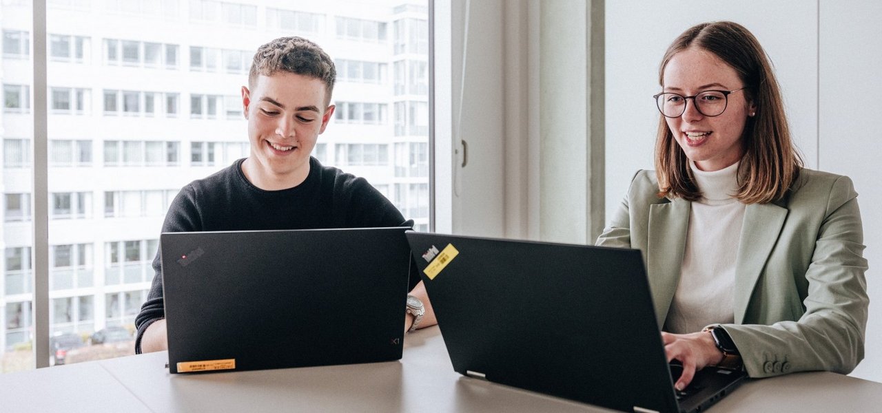 Two doctoral students, a young man and a young woman, sit next to each other and work on their laptops at the Daimler Truck Campus