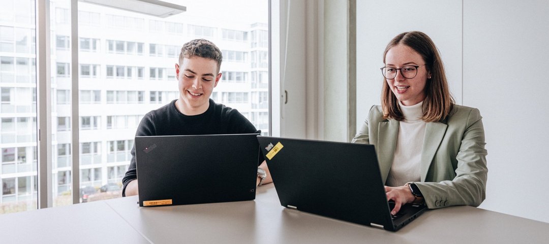 Two doctoral students, a young man and a young woman, sit next to each other and work on their laptops at the Daimler Truck Campus