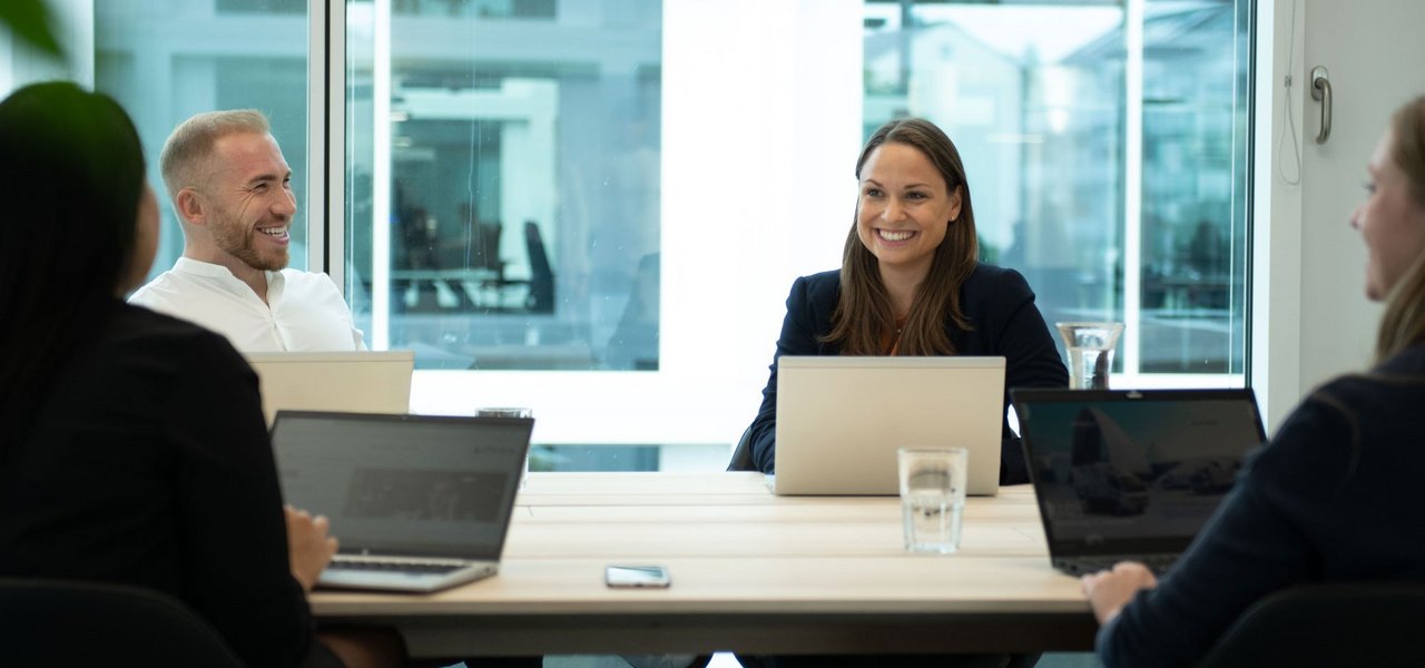 A meeting in a modern office where four employees are chatting in a relaxed atmosphere and taking notes on their laptops.