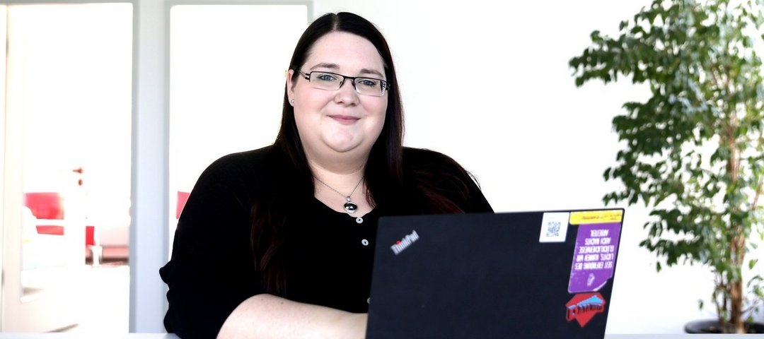 Diana Andrä sits smiling at a desk with a laptop in a bright, modern environment.