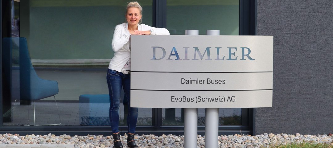 EvoBus PR manager Aylin Flegel leans against a company sign with the Daimler logo in front of a modern office building.