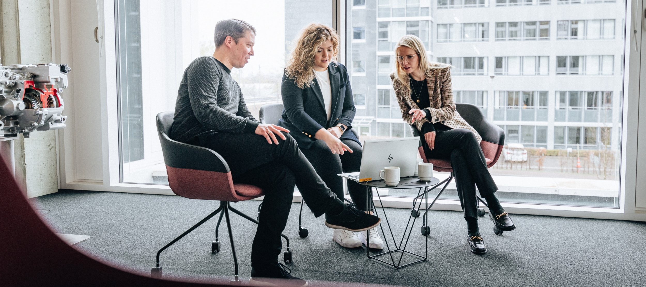Three people are sitting in a brightly lit room on the Daimler Truck Campus, discussing something on a laptop placed on a small table.