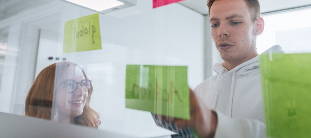 Two young employees stand behind a glass wall with colourful sticky notes, the woman smiles while the man arranges a note.