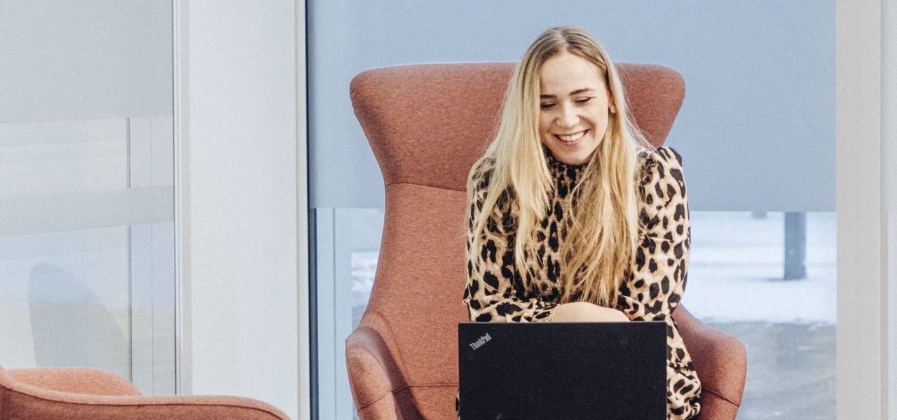 Eine junge Mitarbeiterin sitzt in einem modernen hellen Lounge-Bereich auf einem Sessel und arbeitet an einem Laptop, der auf einem kleinen Tisch vor ihr steht.