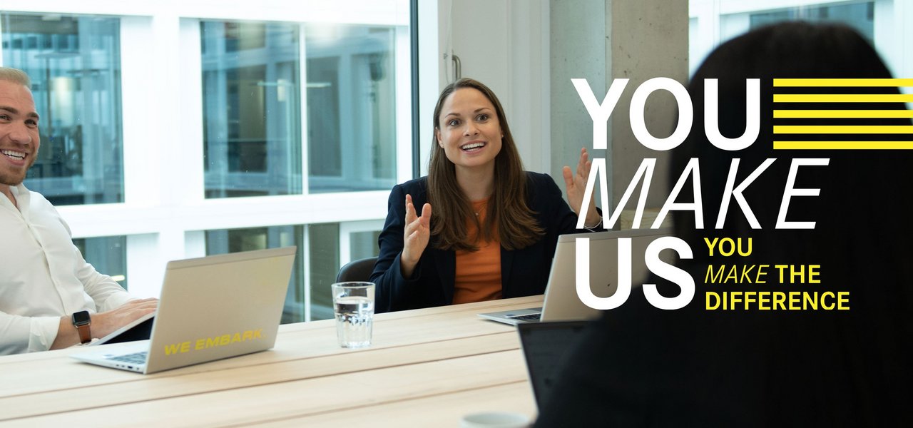 Three colleagues sit across from each other laughing at a meeting table with laptops. Above the picture is the "You Make Us" lettering.