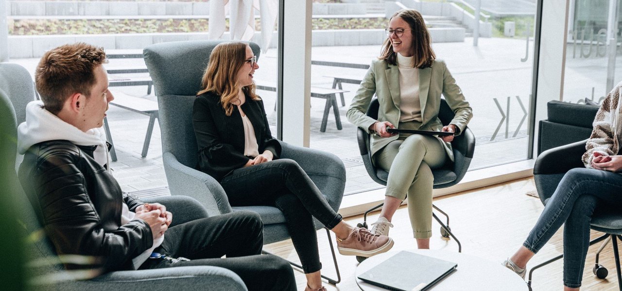 Four students sit on comfortable armchairs in a modern, light-flooded room and chat in a relaxed atmosphere, with an outdoor terrace in the background.