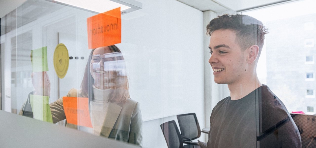 Two young employees stand behind a glass wall with colourful sticky notes, the woman smiles while the man arranges a note.