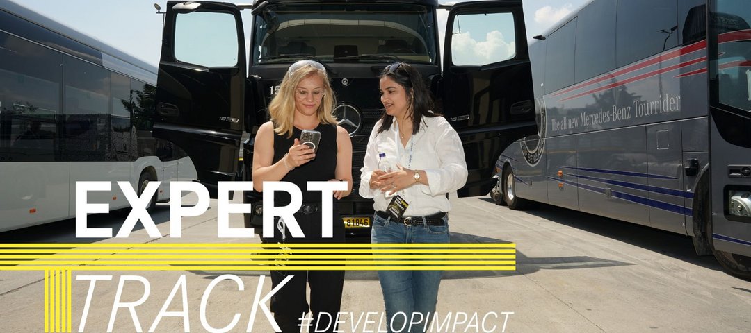 Zwei Daimler Truck Trainee Mitarbeiterinnen schauen auf ein Handy während sie draußen an Daimler Truck Fahrzeugen vorbeilaufen. Auf dem Bild ist der "ExpertTrack" Schriftzug